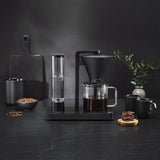 A PERFORMANCE BLACK coffee maker with a transparent water reservoir and a glass carafe filled with coffee. Featuring unique technology for the perfect temperature, it boasts two award badges: one with a laurel wreath indicating "Best i Test Tek.no" and another that reads "Testvinner" with stars and a ribbon symbol.