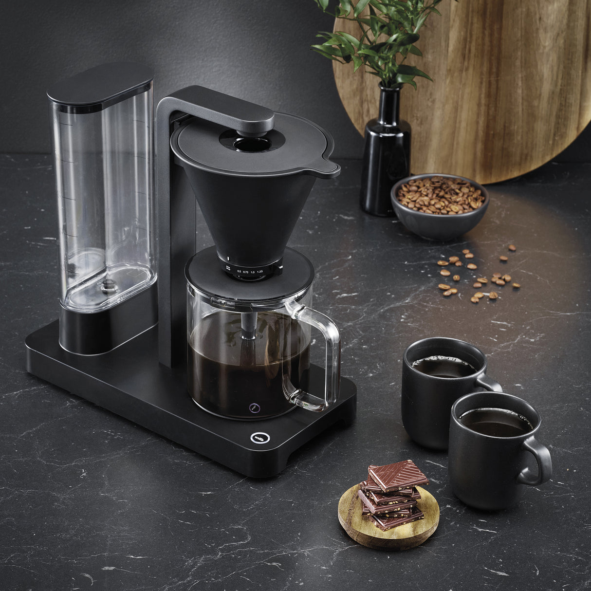 A PERFORMANCE BLACK coffee maker with a transparent water reservoir and a glass carafe filled with coffee. Featuring unique technology for the perfect temperature, it boasts two award badges: one with a laurel wreath indicating "Best i Test Tek.no" and another that reads "Testvinner" with stars and a ribbon symbol.