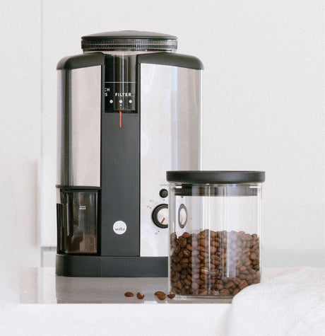 Image of an AeroPress coffee maker with its components separated. From left to right: the gray plunger, the transparent brew chamber with white measurement markings and a silver top, and the black filter cap. The components are arranged on a plain white background, next to a sleek SVART BEAN AND COFFEE CUP W/LID.