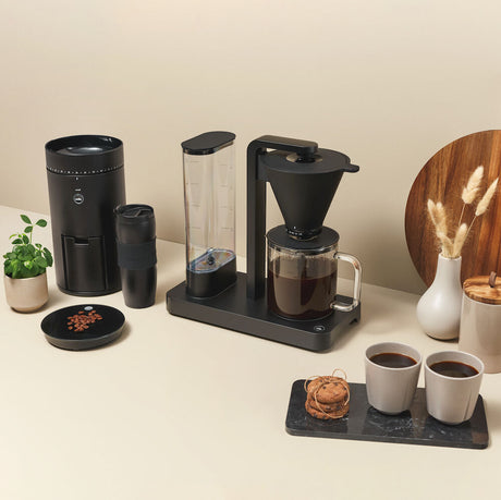 A PERFORMANCE BLACK coffee maker with a transparent water reservoir and a glass carafe filled with coffee. Featuring unique technology for the perfect temperature, it boasts two award badges: one with a laurel wreath indicating "Best i Test Tek.no" and another that reads "Testvinner" with stars and a ribbon symbol.