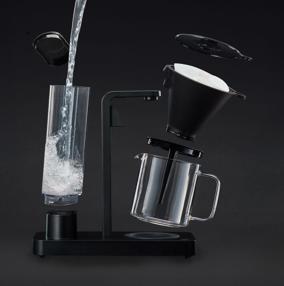 A PERFORMANCE BLACK coffee maker with a transparent water reservoir and a glass carafe filled with coffee. Featuring unique technology for the perfect temperature, it boasts two award badges: one with a laurel wreath indicating "Best i Test Tek.no" and another that reads "Testvinner" with stars and a ribbon symbol.