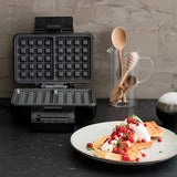 A sleek black GAUFRES BELGIAN with silver accents is pictured against a white background. A gold and black badge with the text "TESTVINNER" is displayed in the top left corner, indicating it has been awarded a test winner. The waffle maker features adjustable temperature and double release coating for perfect waffles every time.